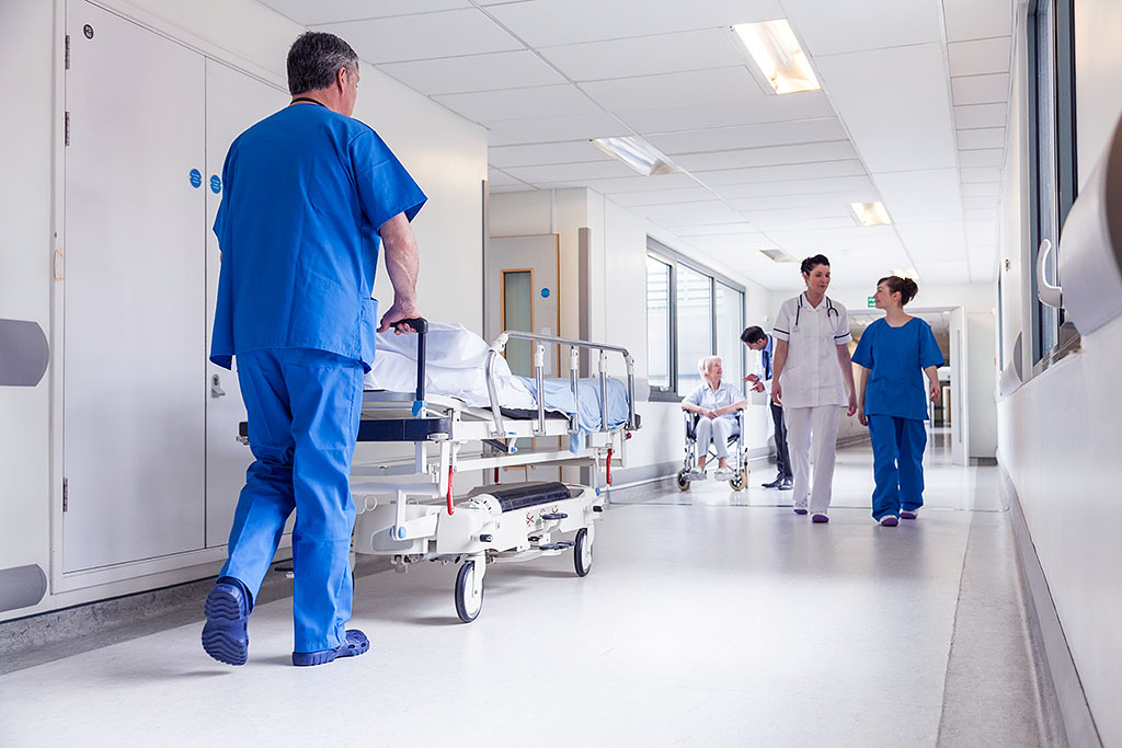 Bright hospital hallway with medical staff in blue scrubs pushing bed, patients in wheelchairs, and large windows along corridor.
