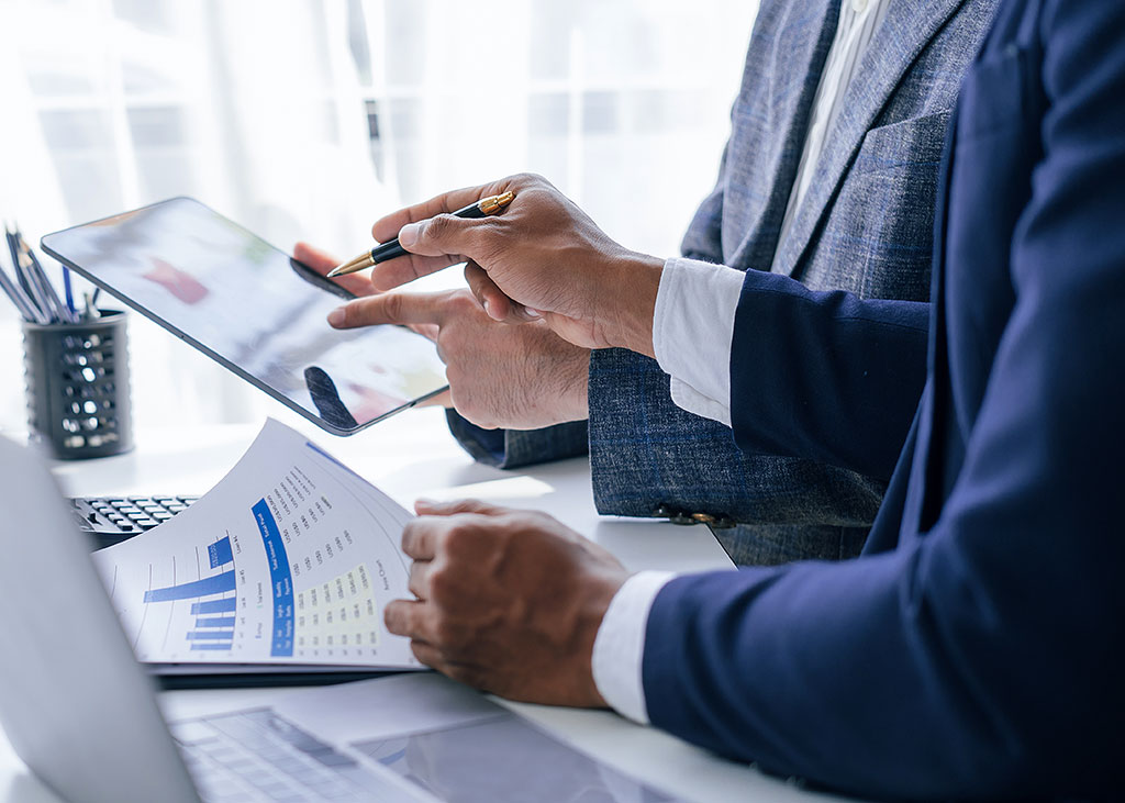 Two business professionals reviewing financial documents and tablet data during a collaborative meeting at a light-toned office table.