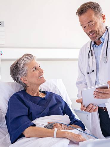 Doctor in white coat consulting smiling female patient in bright hospital room with medical equipment and digital tablet.