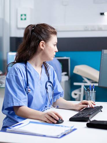Female medical professional in light-blue scrubs reviewing data on computer in organized, modern clinic with teal walls and medical supplies.