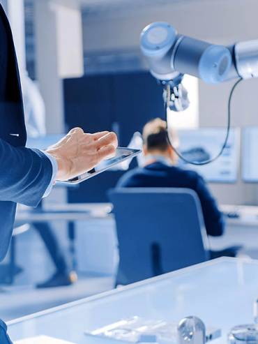 Person in dark suit holding tablet in modern industrial workspace with robotic arm, technical monitors, and assembly components.