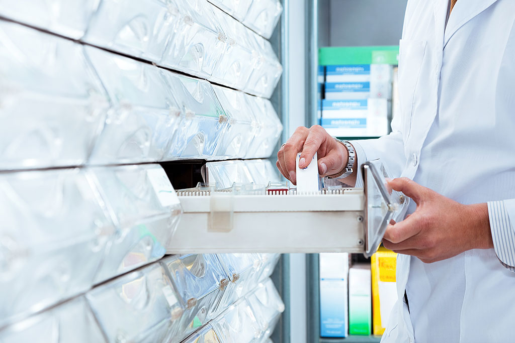 Pharmacist in white coat retrieving prescription from organized wall-mounted storage with transparent compartments in bright pharmacy.