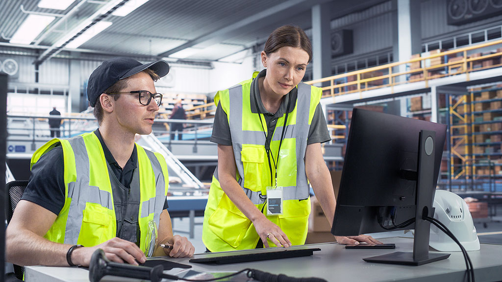 Two individuals focused on a computer screen inside a large industrial warehouse, emphasizing digital workflow in logistics.
