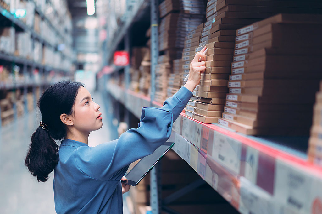 Young woman in light-blue shirt checking inventory on shelves in bright, organized warehouse using tablet and stylus.