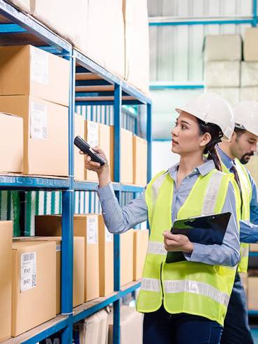 Woman in yellow shirt scanning barcodes on boxes in bright warehouse, performing inventory stocktaking with handheld scanner.